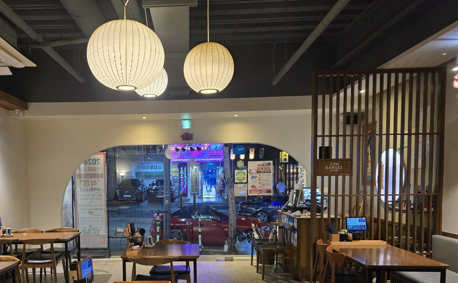 칸세이스시 상무본점 매장 — 리모델링 뒤 홀 전경. 어두운 노출 천장에 한지 구형 등이 매달려 있고, 가운데 아치형 유리 입구 너머로 밤거리 불빛이 보인다. 오른쪽에는 KANSEI 명판을 건 월넛 세로 살창 칸막이, 앞쪽에는 원목 테이블과 의자가 놓여 있다.