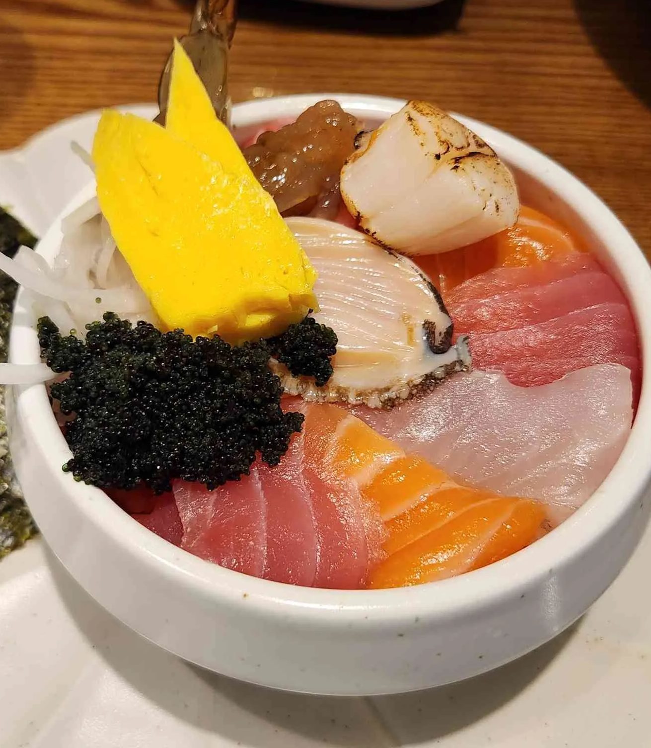 칸세이스시 활짝 핀 카이센동 — 꽃 모양 흰 받침 접시 위 흰 그릇에 담긴 카이센동. 참치, 연어, 흰살생선, 전복 조각, 겉을 그을린 관자, 계란말이, 검은 날치알을 돌려 담고 새우 꼬리를 꽂았으며, 옆에 김이 놓여 있다.