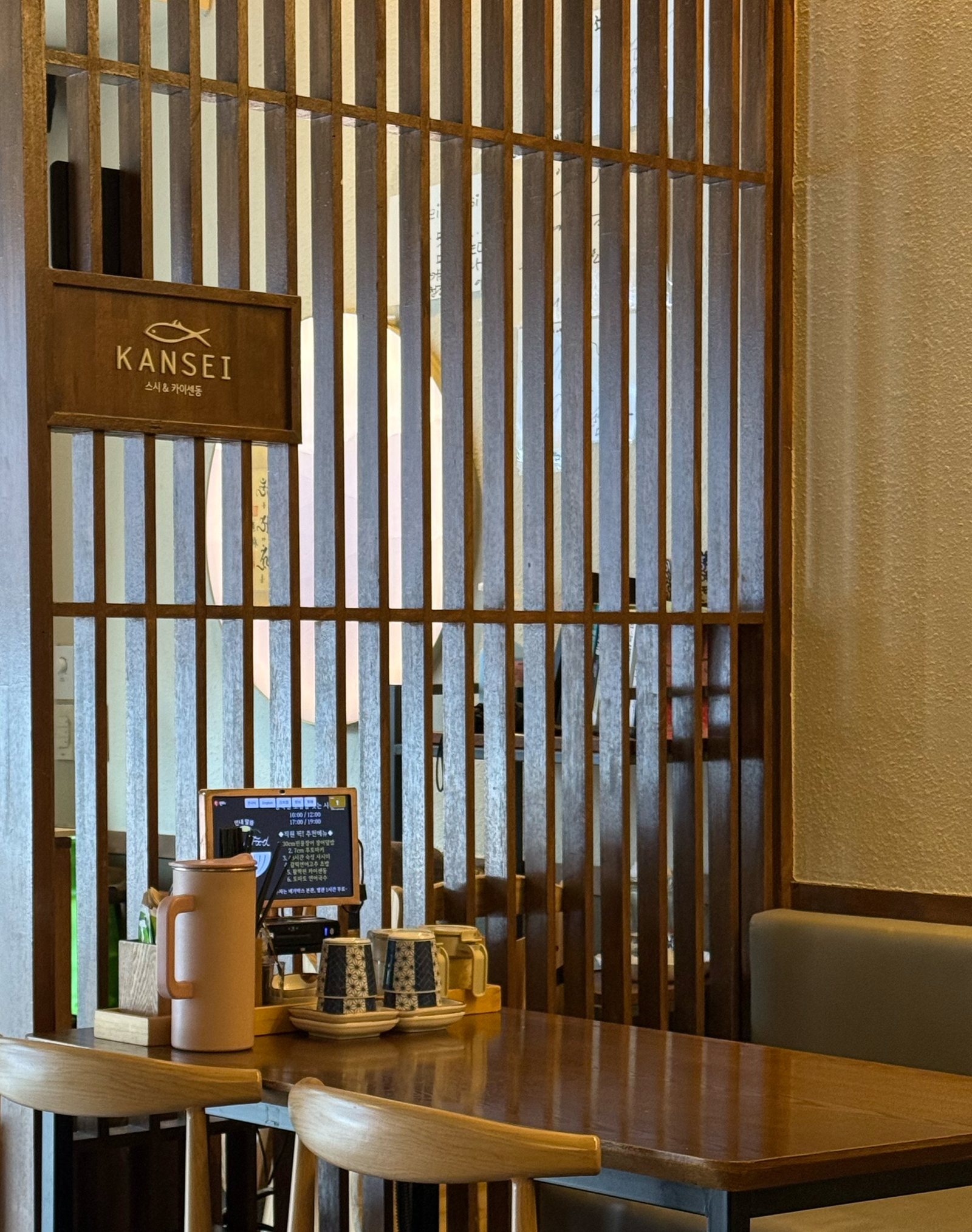 칸세이스시 상무본점 매장 — 월넛 세로 살창 칸막이에 건 KANSEI 물고기 로고 나무 명판(스시 & 카이센동). 살창 사이로 둥근 빛이 비치고, 앞에 원목 테이블과 물병, 주문용 태블릿이 놓여 있다.