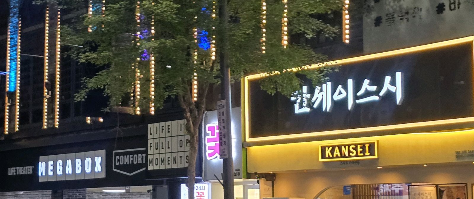 칸세이스시 상무본점 외관 — 밤의 메가박스 본관 앞 거리. 왼쪽에 MEGABOX·LIFE THEATER·COMFORT 간판과 전구를 두른 가로수, 오른쪽에 불 켜진 '칸세이스시' 간판과 노란 KANSEI 박스 간판이 나란히 보인다.