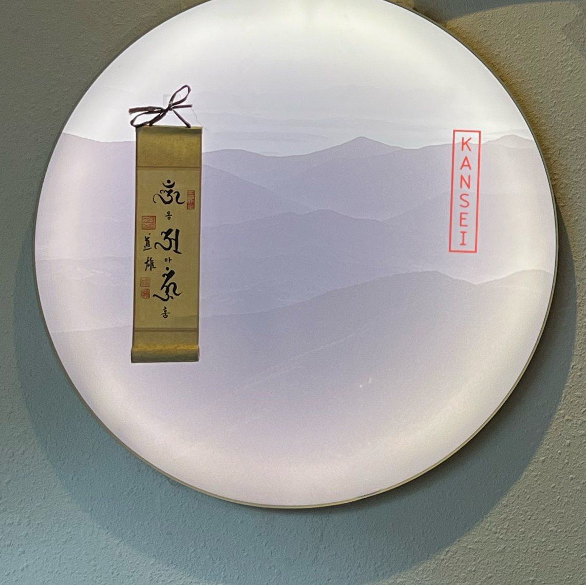 칸세이스시 상무본점 매장 — 안개 낀 산 능선이 옅게 그려진 둥근 조명 패널. 가운데에 붓글씨 족자가 걸려 있고 오른쪽에 붉은 세로 KANSEI 낙관이 있다.