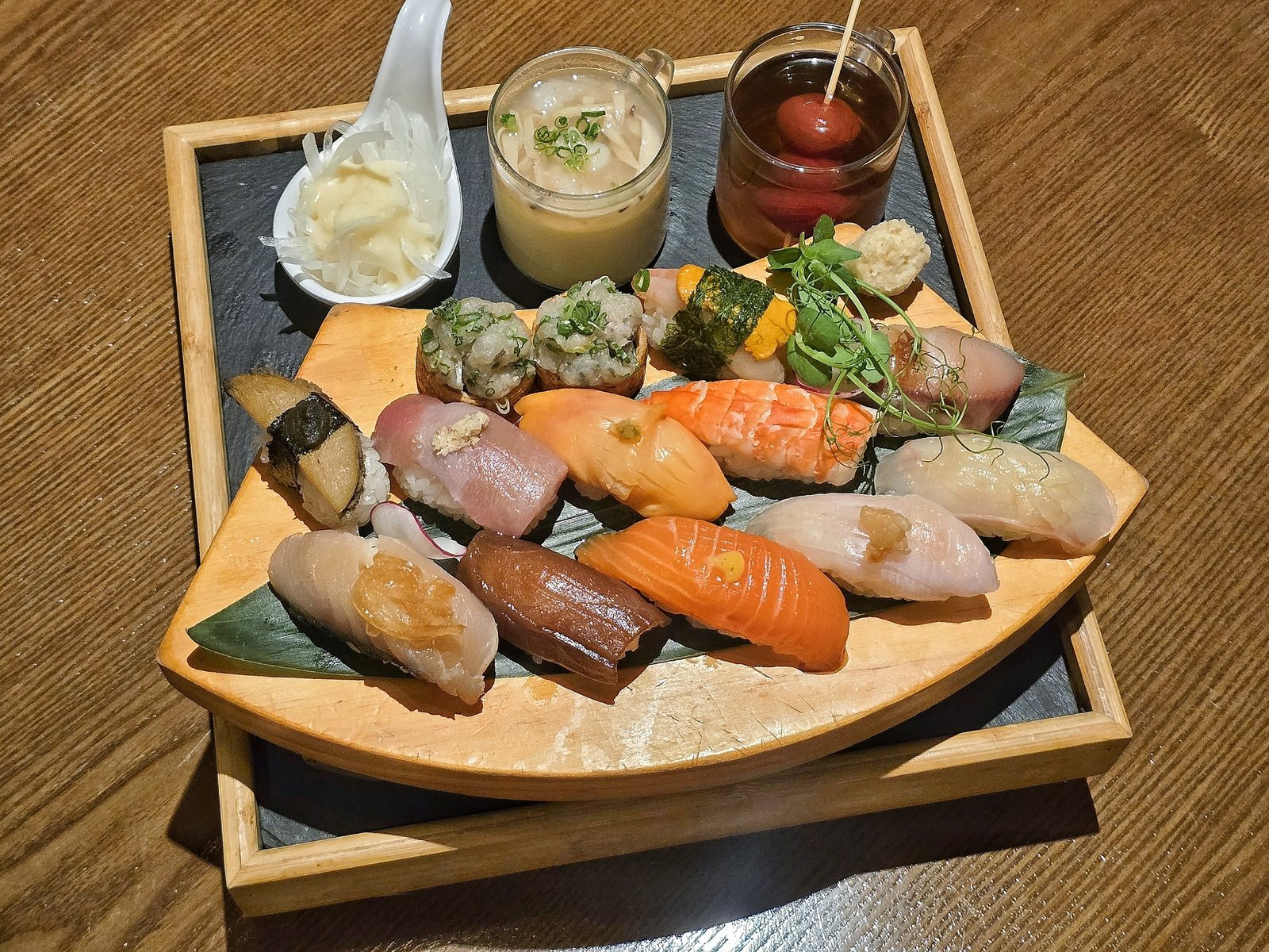 칸세이스시 캐주얼 초밥 오마카세 — 원목 테이블 위 검은 석판 쟁반에 부채꼴 원목 판을 올리고 초밥 한 판을 담았다. 연어, 새우, 참치, 흰살생선 초밥과 김을 두른 군함, 파를 올린 초밥이 있고, 뒤로 계란찜과 토마토 절임 잔, 초생강을 담은 숟가락이 놓여 있다.