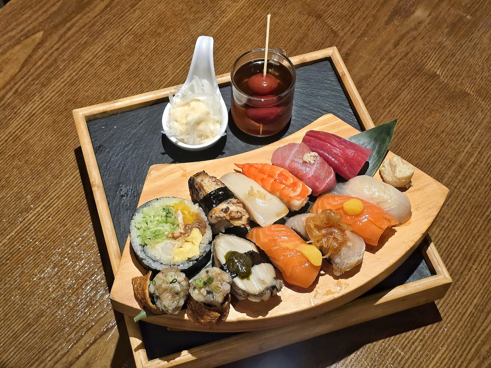 칸세이스시 모둠 초밥 한 판과 후토마키 — 원목 테이블 위 검은 석판 쟁반에 올린 원목 판. 오이·계란말이·게살을 흰 밥으로 싼 두툼한 후토마키 한 조각과 새우·참치·연어·오징어·붕장어·전복 초밥과 유부초밥이 놓여 있고, 뒤로 초생강 숟가락과 토마토 절임 잔이 보인다.