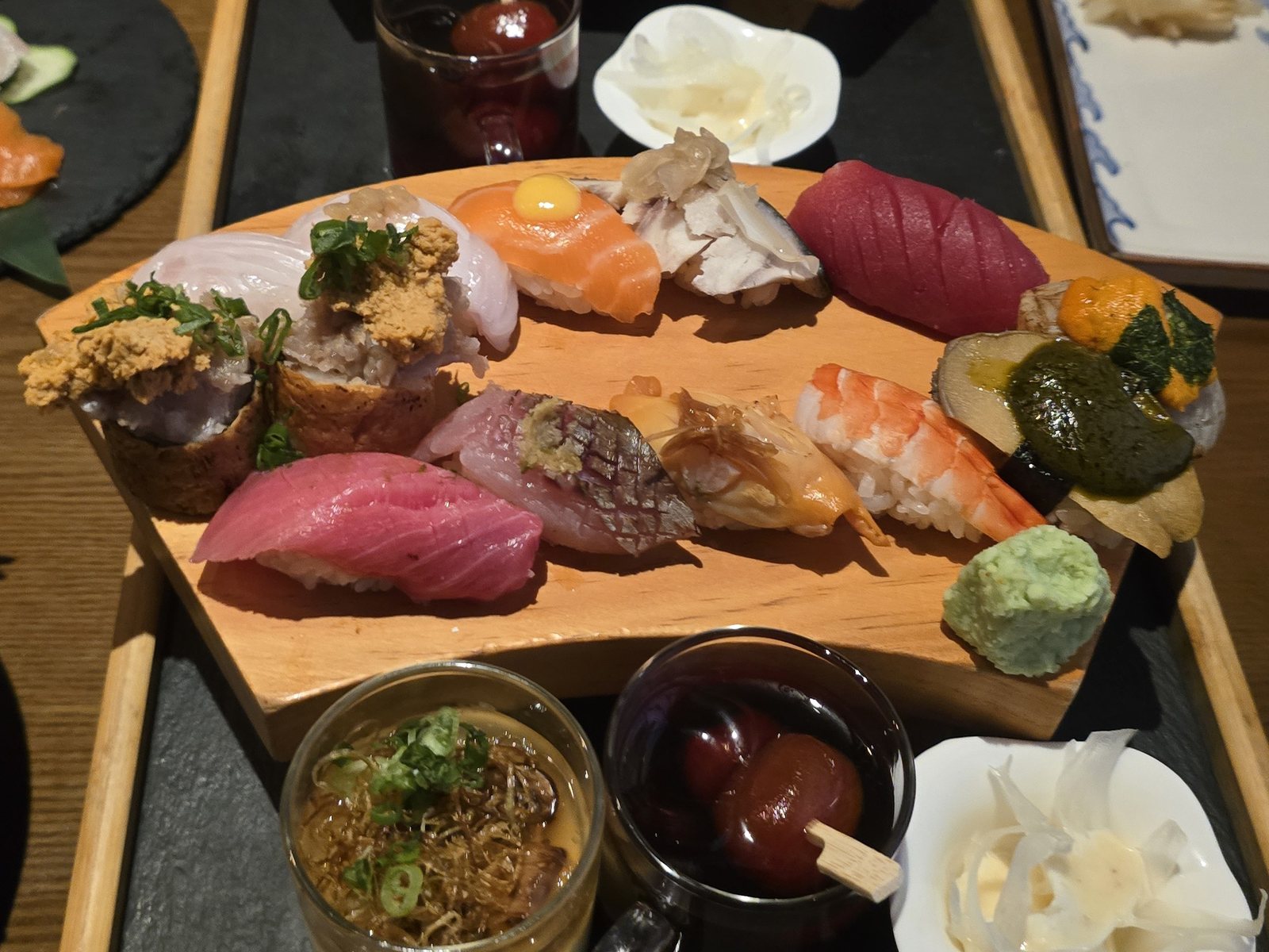 칸세이스시 모둠 초밥 한 판 (참치 뱃살·전갱이·성게알 군함) — 검은 석판 쟁반 위 원목 판에 담긴 초밥. 참치 붉은살과 뱃살, 칼집 낸 전갱이, 메추리알 노른자를 올린 연어, 새우 초밥과 토핑을 얹은 유부초밥이 있고, 앞쪽에 계란찜과 토마토 절임 잔, 초생강이 놓여 있다.