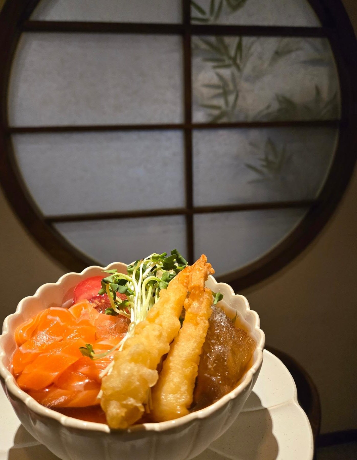 칸세이스시 토마토 연어국수 — 대나무 그림자가 비치는 원형 격자 창 앞, 꽃 모양 흰 그릇에 담긴 연어국수. 연어회와 방울토마토, 새우튀김 두 개, 무순을 올렸고 얼음 조각이 든 국물이 보인다.