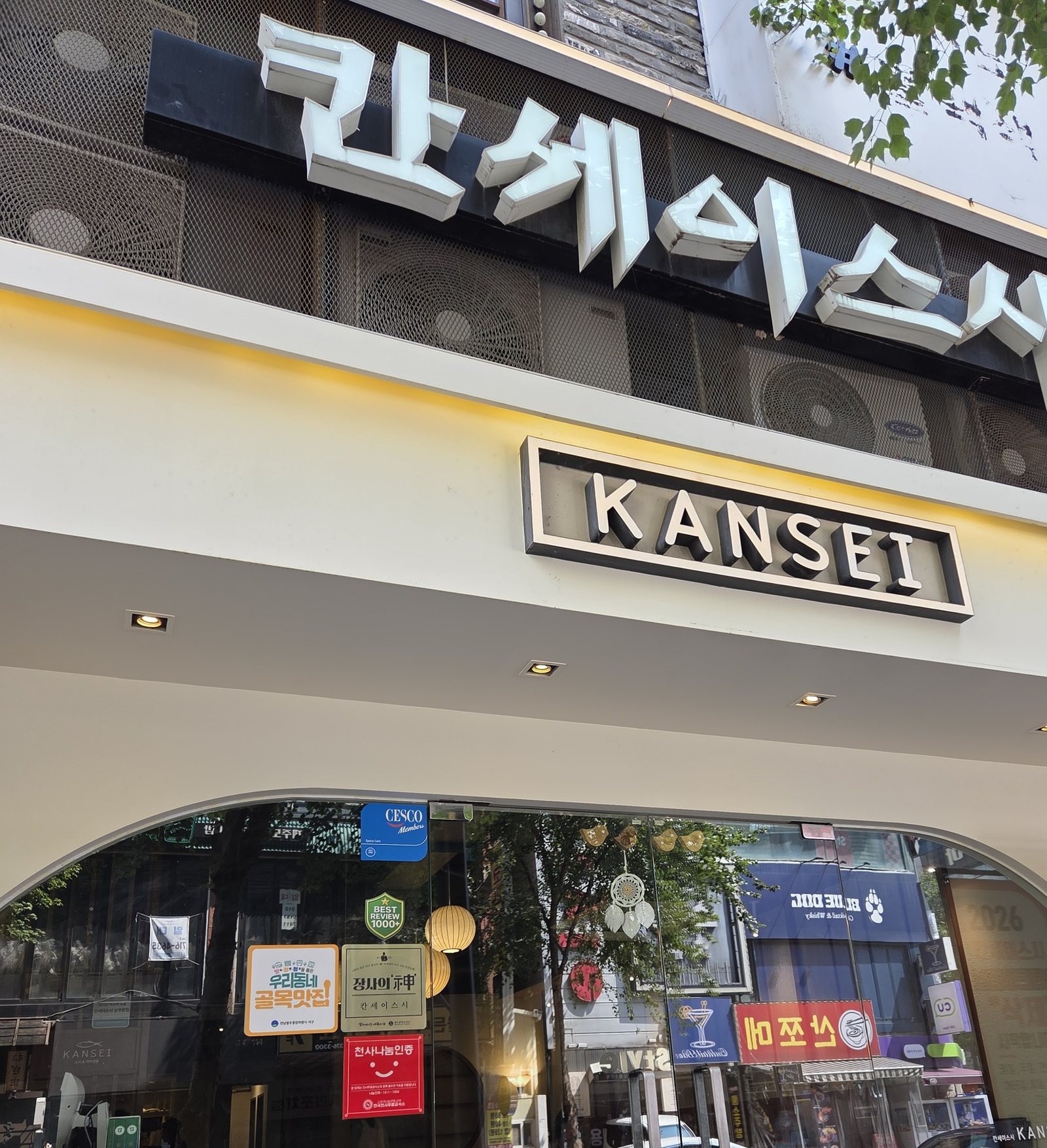 칸세이스시 상무본점 외관 — 낮의 매장 정면을 왼쪽 아래에서 올려다본 모습. 흰 입체 글자 '칸세이스시' 간판과 KANSEI 박스 간판, 노란 LED 띠, 캐노피 아래 아치형 유리 입구가 보이고 유리에 인증 스티커와 거리 풍경이 비친다.
