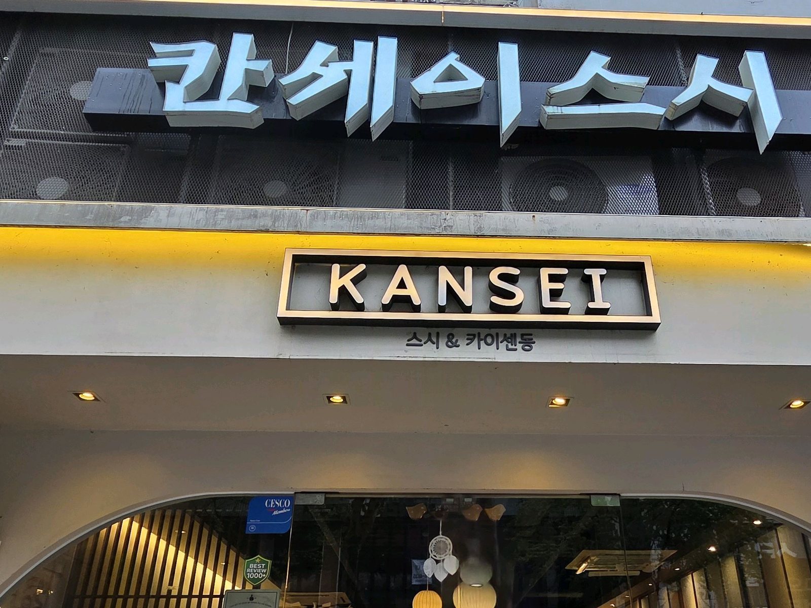칸세이스시 상무본점 외관 — 해 질 녘 매장 정면을 올려다본 모습. 짙은 타공 패널 위 흰 입체 글자 '칸세이스시' 간판, 노란 LED 띠 아래 'KANSEI 스시 & 카이센동' 박스 간판, 불이 켜진 아치형 유리 입구 안쪽의 한지 등과 세로 살창이 보인다.