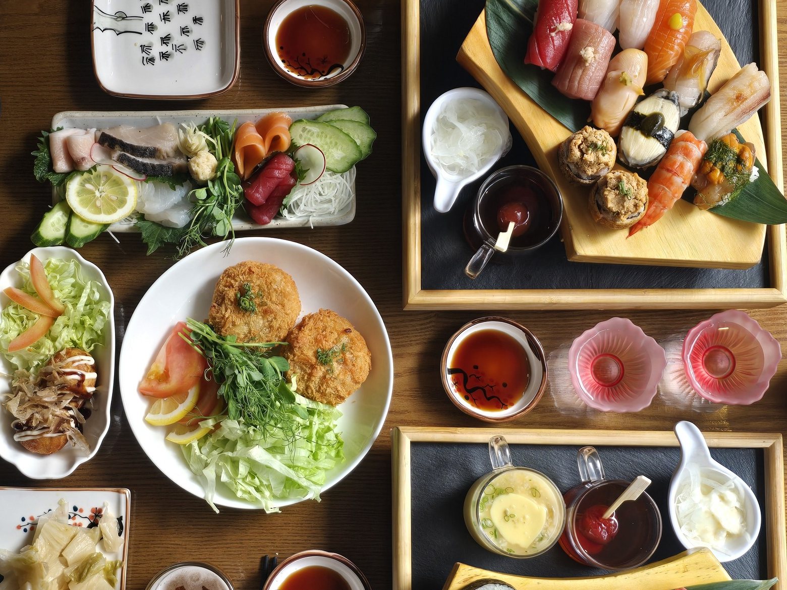 칸세이스시 2인 상차림 (초밥 두 판·사시미·멘치카츠 샐러드) — 위에서 내려다본 두 사람 상차림. 오른쪽 위 부채꼴 나무 판에 담긴 초밥 한 판, 겉을 그을린 생선·연어·참치를 담은 사시미 접시, 멘치카츠와 양배추 샐러드, 계란찜, 분홍 꽃잔, 간장 종지가 원목 테이블에 놓여 있고 오른쪽 아래에 두 번째 초밥 판이 보인다.