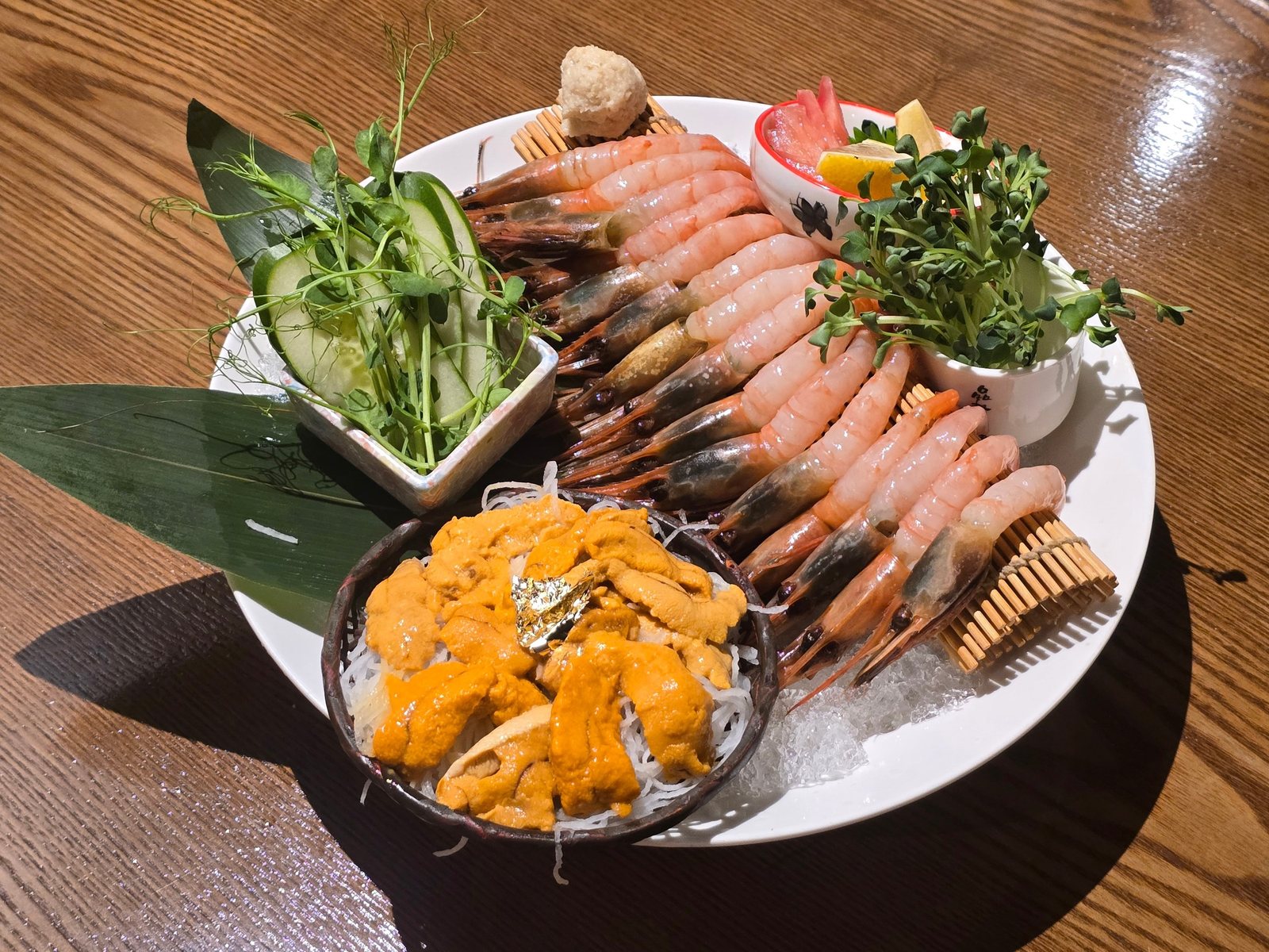 칸세이스시 우니 단새우 사시미 — 원목 테이블 위 흰 원형 접시에 대발을 깔고 부채꼴로 늘어놓은 단새우, 채 썬 무 위에 금박을 얹은 성게알 한 그릇, 오이와 완두순을 담은 사각 그릇, 무순을 꽂은 컵과 큰 잎 장식이 놓여 있다.